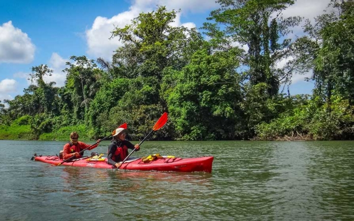 A red kayak is paddled by two outward bound students on calm water. The shore is lined with green trees and the sky is blue with white clouds.
